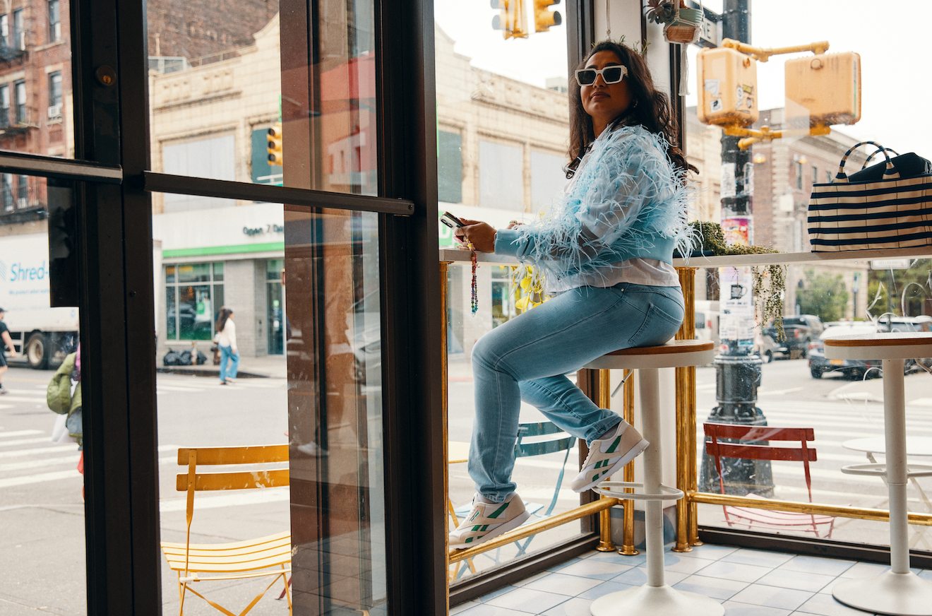 woman sitting on a high chair in the window of a cafe, wearing a blue fluffy jacket and reebok veefriends shoes